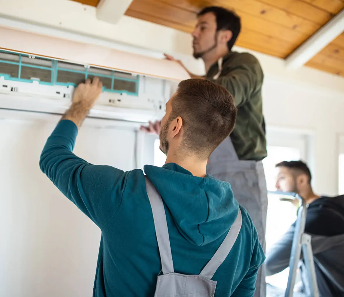 Two men working on an air conditioner in a room with a wall in the background