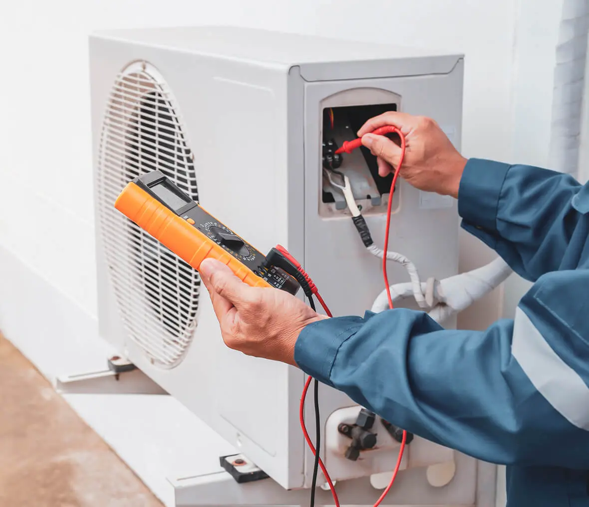 Man in a uniform holding a multimeter and checking an air conditioner, with wires connected to the device