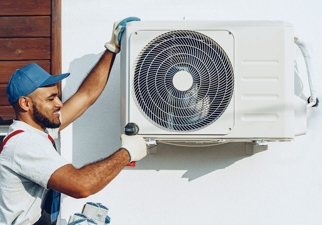 Man wearing a cap and gloves, holding a tool and fixing an air conditioner on the wall of a house